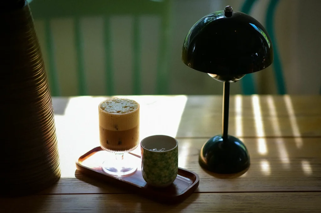 A cozy minimalist cafe table scene featuring an iced latte with foam in a ribbed glass, placed on a small tray beside a patterned ceramic cup. A black dome table lamp sits on the right, casting soft shadows across the wooden tabletop, while warm sunlight streams through the background, creating a calm and moody atmosphere.