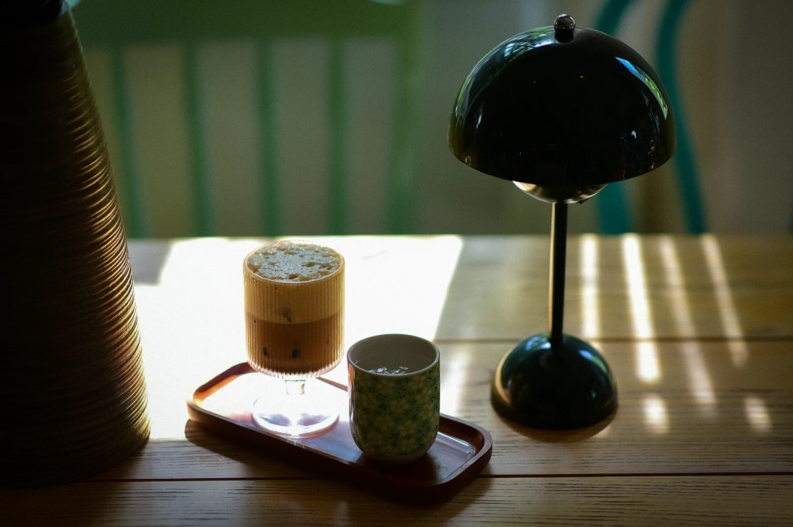 A cozy minimalist cafe table scene featuring an iced latte with foam in a ribbed glass, placed on a small tray beside a patterned ceramic cup. A black dome table lamp sits on the right, casting soft shadows across the wooden tabletop, while warm sunlight streams through the background, creating a calm and moody atmosphere.