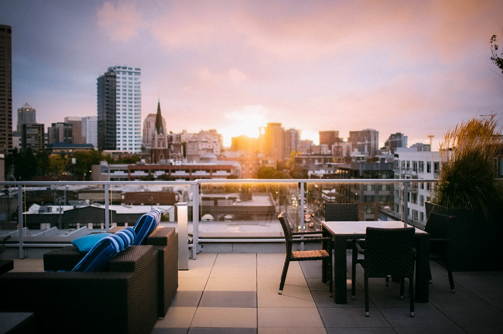 Modern rooftop dining terrace overlooking a city skyline at sunset, featuring outdoor lounge seating, dining tables, glass railings, and warm golden-hour views across urban buildings.