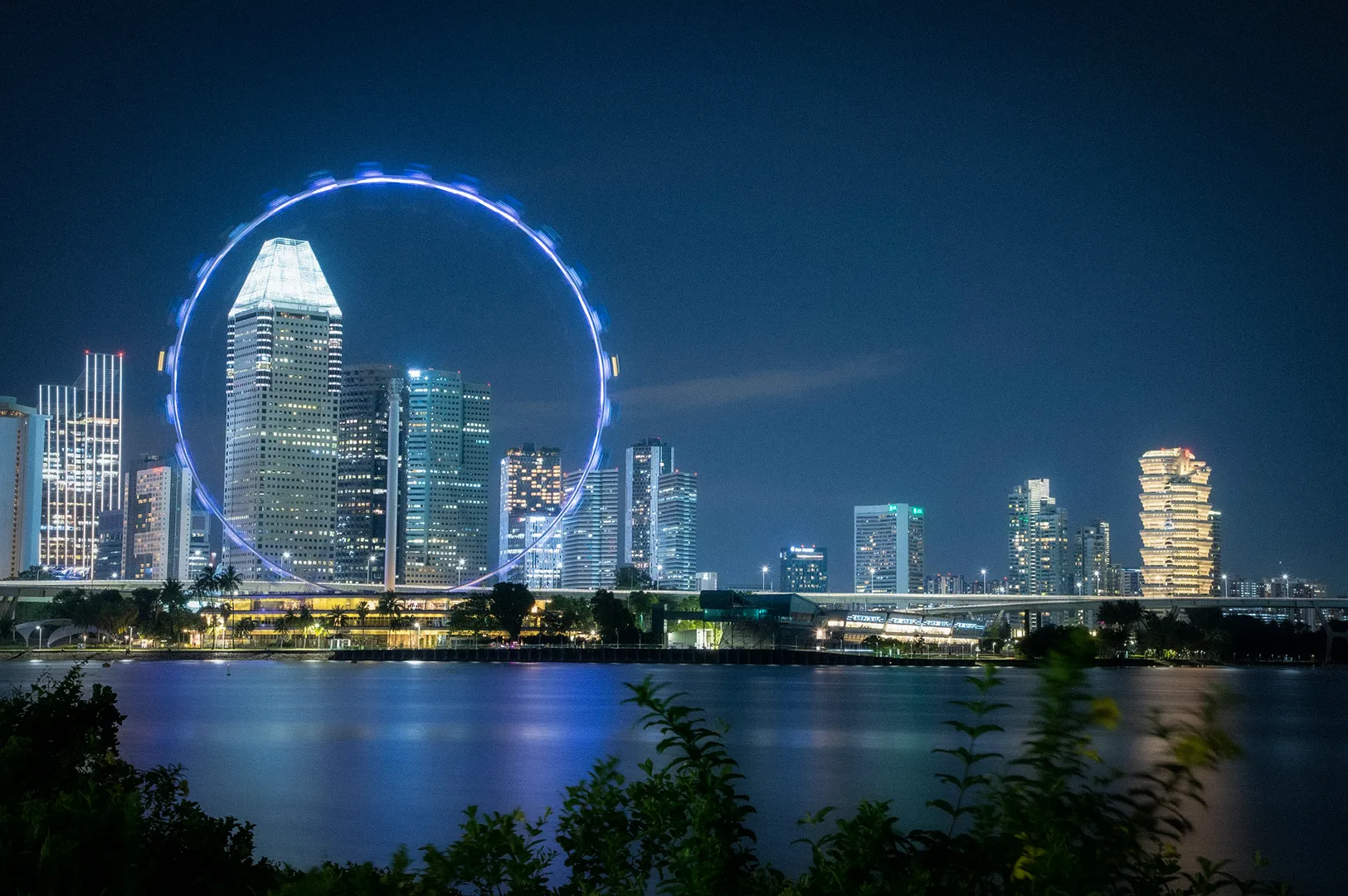 Night view of Singapore’s skyline featuring the Singapore Flyer illuminated against the cityscape. Modern skyscrapers and waterfront developments reflect on the calm waters of Marina Bay, creating a vibrant urban scene with glowing lights and architectural landmarks.