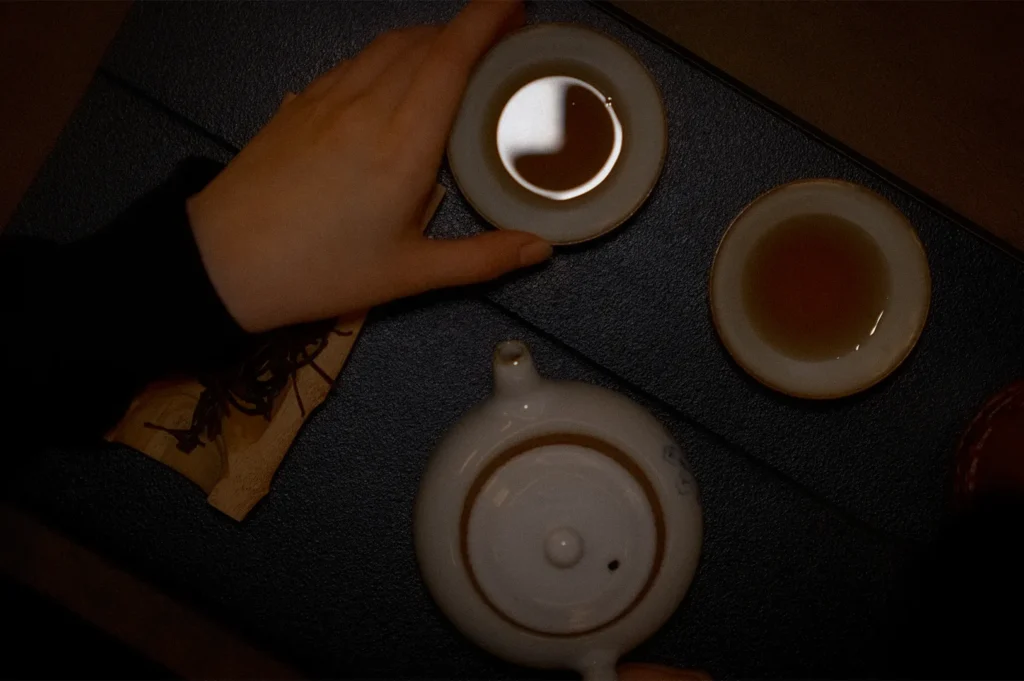 Overhead view of a traditional tea set with ceramic teapot and teacups during a tea tasting experience at Tea Room by Ki-Setsu in Orchard Plaza, Singapore. A guest's hand reaches for a cup of brewed tea on a dark textured tea table.