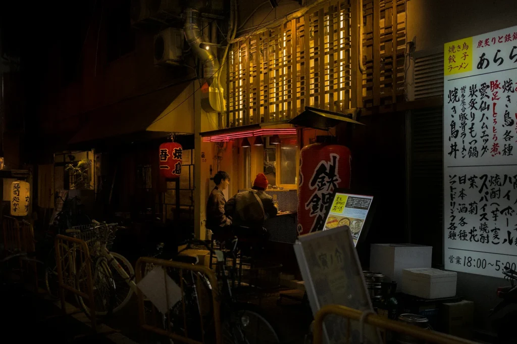 Moody nighttime street scene of a traditional Japanese izakaya alley with glowing lanterns, handwritten menu signs, bicycles, and customers dining at a small open-front yakitori bar in Tokyo, Japan