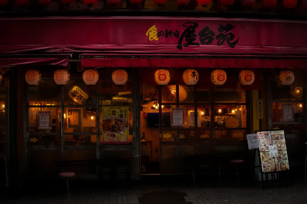 Traditional Japanese izakaya restaurant exterior illuminated by paper lanterns at night, featuring a cozy storefront, menu display, warm interior lighting, and authentic Japanese dining atmosphere.