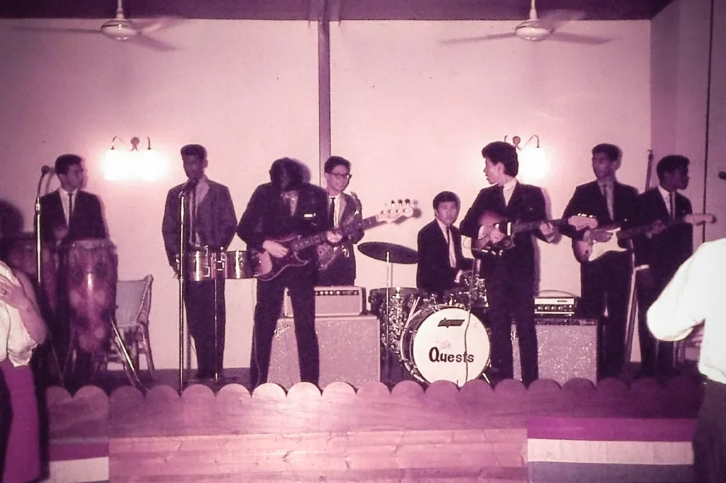 Vintage photograph of The Quests performing live on stage in Singapore during the 1960s. The influential Singaporean rock band is seen playing guitars and drums before a dance floor audience, capturing the energy of the country's early pop and rock music scene.
