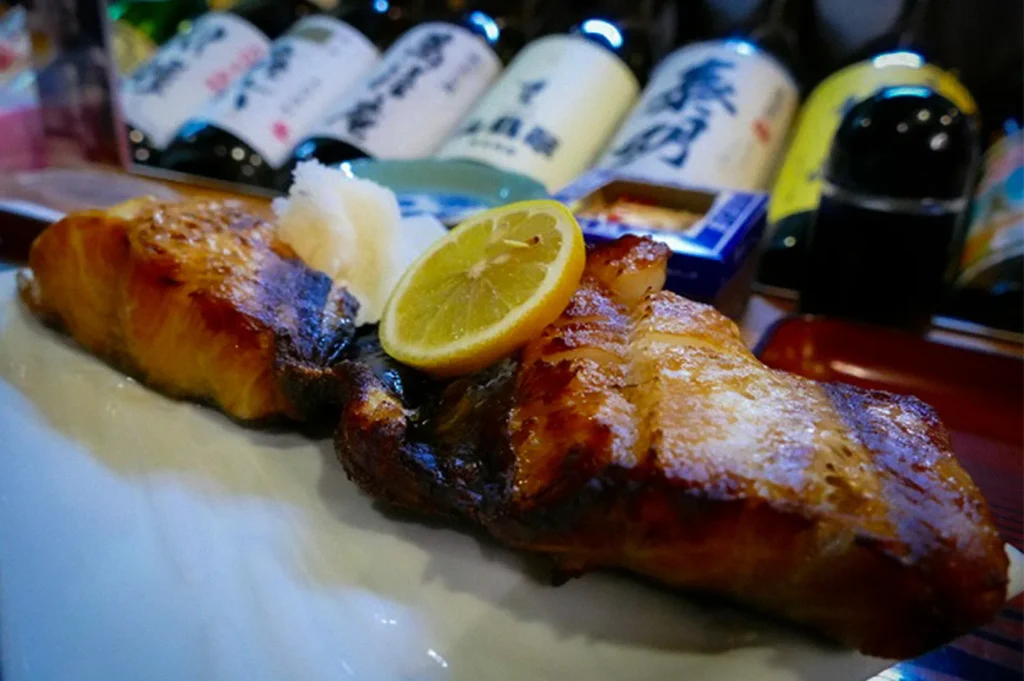 Grilled salmon fillet served on a plate with lemon and grated daikon at a Japanese izakaya, with sake bottles displayed in the background highlighting traditional Japanese pub dining.