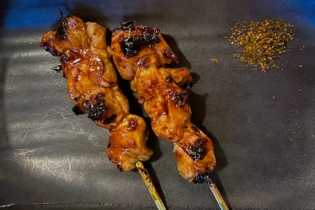 Japanese yakitori chicken skewers glazed with savory sauce and lightly charred over the grill, served on a black plate with Japanese spice seasoning at a traditional izakaya restaurant.