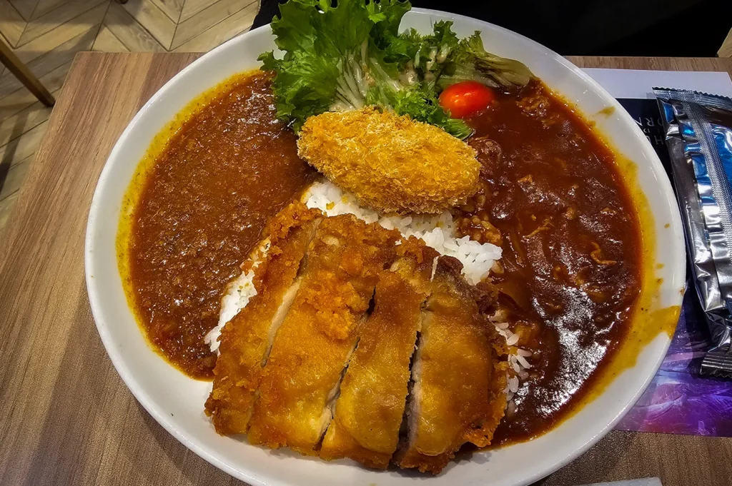 Japanese curry rice at Japan Rail Cafe Singapore featuring crispy chicken katsu, a golden breaded croquette, fresh salad, and rich curry sauces served over steamed rice in a large bowl.