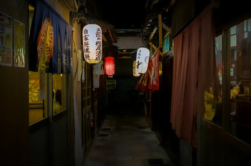 Japanese-style izakaya alley in Singapore with glowing paper lanterns, cozy restaurant entrances, traditional noren curtains, and an intimate nighttime dining atmosphere.
