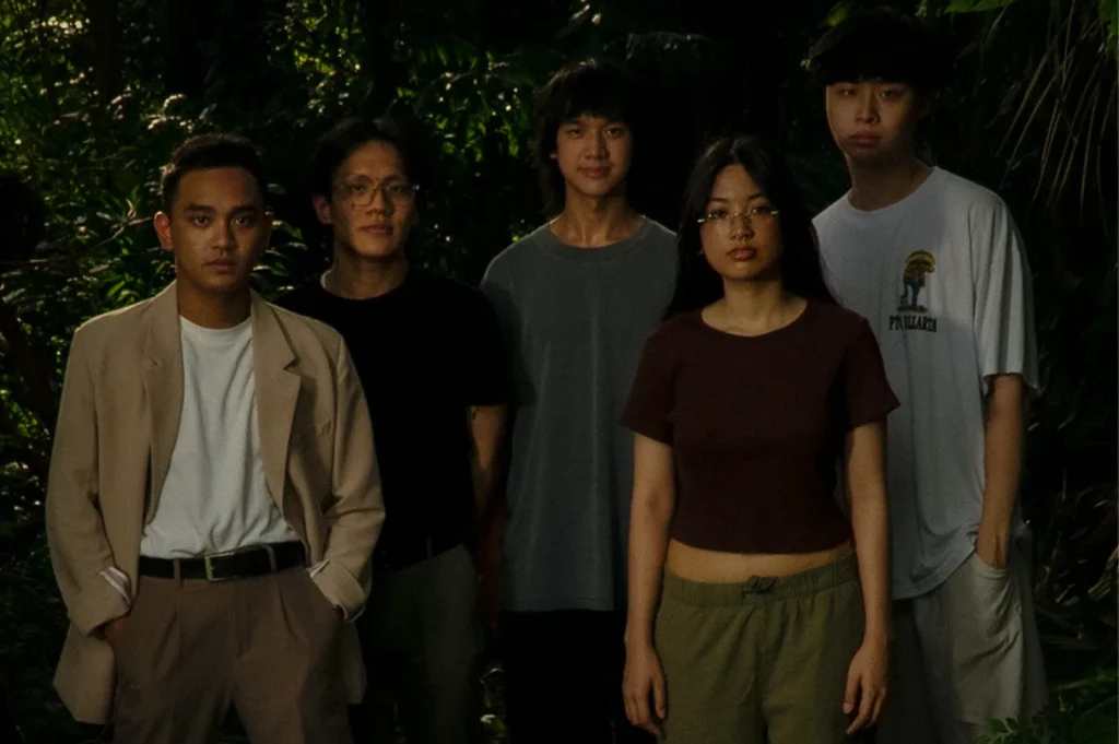 Promotional portrait of Woes, a Singapore indie band, posing together in a lush tropical garden setting. The five-member group stands among dense greenery, reflecting the contemporary aesthetic and creative identity of Singapore's independent music scene.