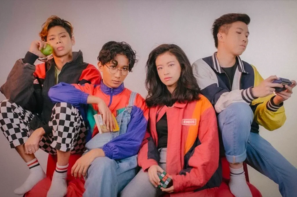 Promotional portrait of Disco Hue, a Singapore indie pop band, featuring four members seated against a clean studio backdrop. The group wears colorful retro-inspired streetwear, reflecting the vibrant, youthful energy of Singapore's contemporary music scene.