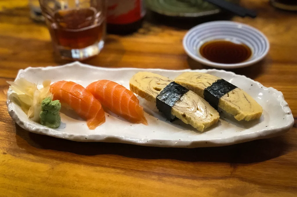 Fresh salmon nigiri and tamago sushi served on a ceramic plate with wasabi, pickled ginger, and soy sauce at a traditional Japanese izakaya restaurant.