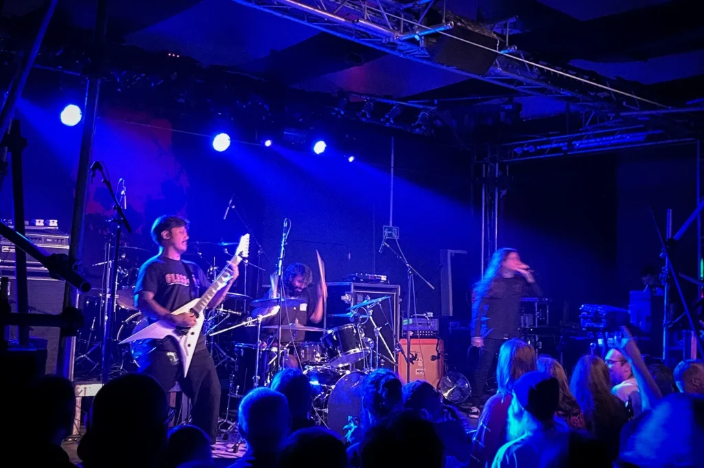 Wormrot performing live on stage at a packed concert venue, with the Singapore grindcore band's vocalist, guitarist, and drummer delivering an intense performance under blue stage lighting. Audience members gather near the stage as amplifiers, drum kits, and professional concert equipment create an energetic underground metal music atmosphere.