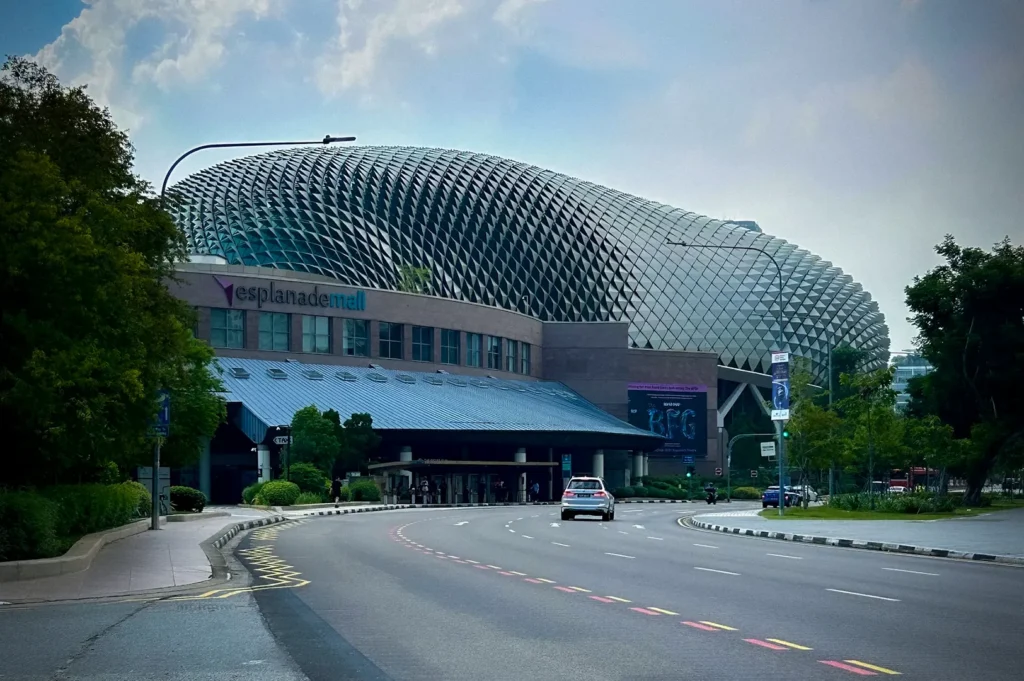 Exterior view of the Esplanade – Theatres on the Bay in Singapore, featuring its distinctive durian-inspired architectural facade beside Esplanade Mall. The iconic performing arts center stands along a tree-lined road in the Marina Bay area under a bright sky, showcasing one of Singapore’s most recognizable cultural landmarks.