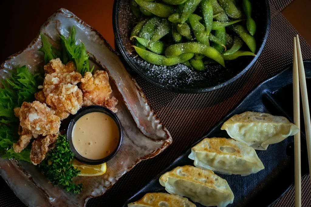 Japanese izakaya appetizers featuring pan-fried gyoza dumplings, crispy karaage fried chicken with dipping sauce, and salted edamame served on rustic ceramic plates with chopsticks.