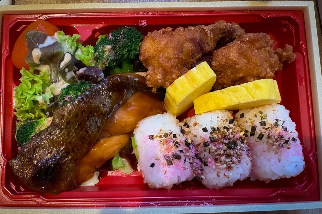Japanese bento box meal at Japan Rail Cafe Singapore featuring grilled salmon, karaage fried chicken, tamagoyaki Japanese omelette, seasoned rice balls, fresh salad, and vegetables served in a traditional bento-style tray.