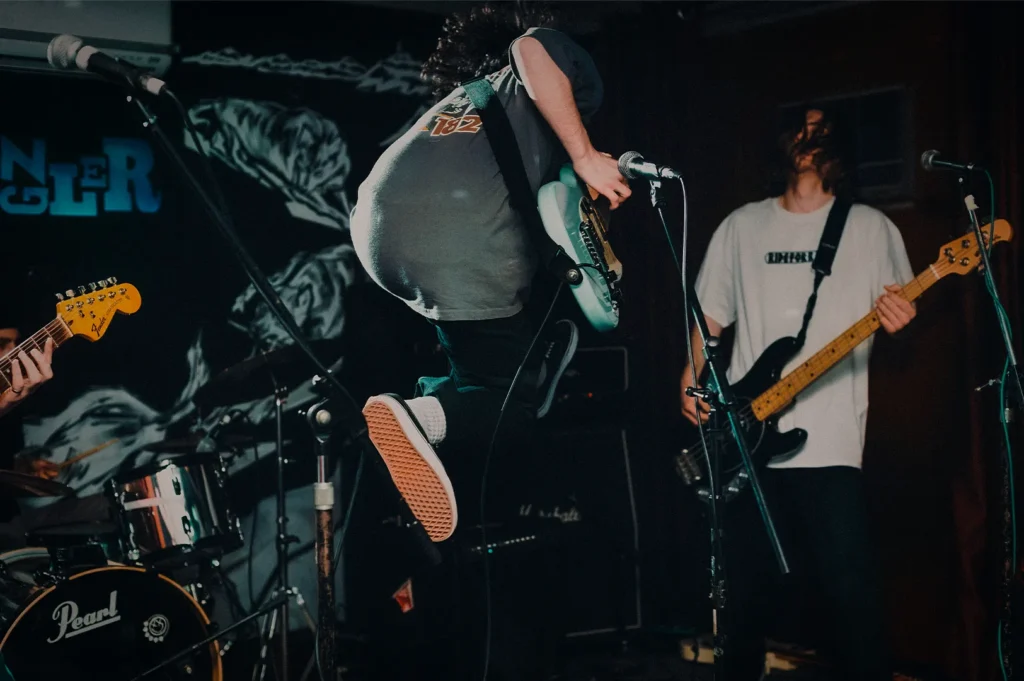 Independent rock band performing live on stage during an energetic concert, featuring musicians playing electric guitar, bass guitar, and drums under stage lighting. A guitarist jumps mid-performance while band members perform in a small music venue surrounded by microphones, amplifiers, and professional live sound equipment, capturing the excitement of an indie music show.