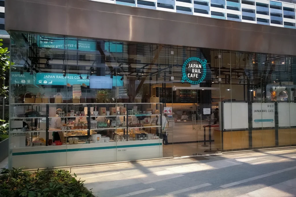 Exterior view of Japan Rail Cafe Singapore featuring a modern glass storefront, Japanese retail products, Japan Rail Pass counter services, and cafe dining area visible through the floor-to-ceiling windows.