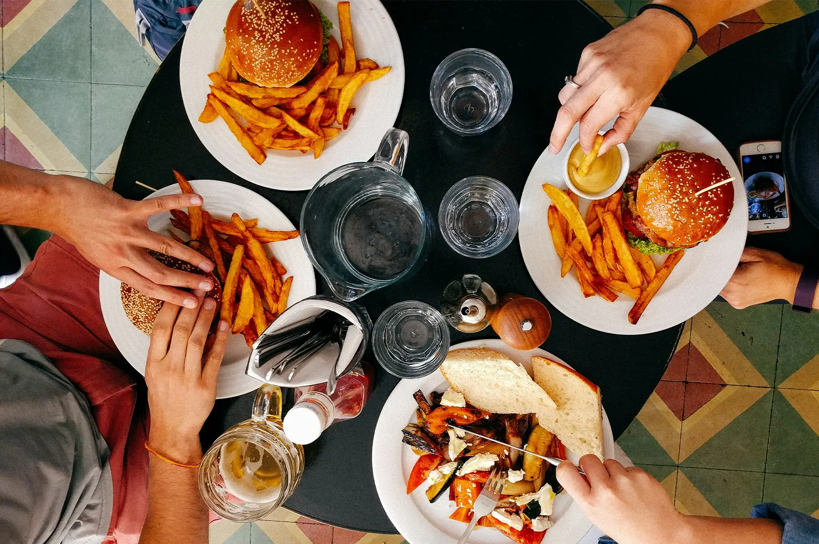 Friends sharing burgers, fries, and roasted vegetables around a dining table, captured from an overhead view in a casual restaurant setting.