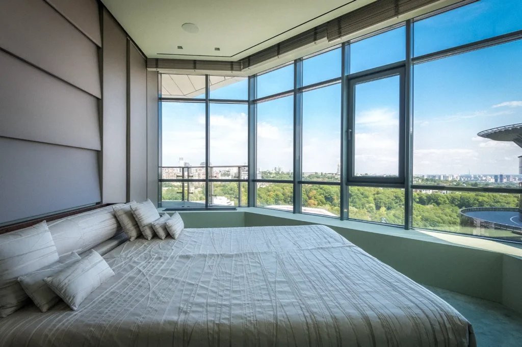 Modern luxury hotel room with a king-size bed, floor-to-ceiling windows, and panoramic city skyline views, featuring a bright contemporary interior and natural daylight.
