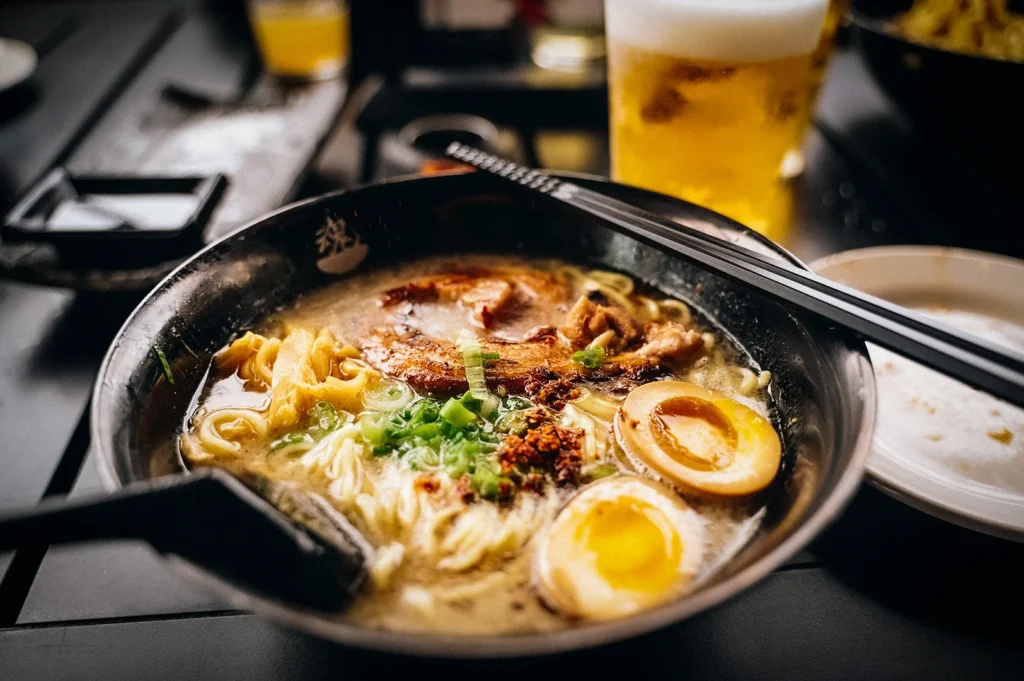 Japanese tonkotsu ramen with chashu pork, soft-boiled egg, green onions, chili flakes, and noodles served in a rich broth with beer in the background.