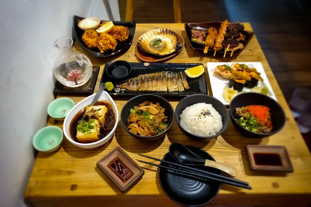 Japanese feast at Shukuu Japanese Izakaya Singapore featuring grilled mackerel, yakitori skewers, karaage, sushi, tofu, rice bowls, sake, and assorted izakaya dishes on a wooden table.