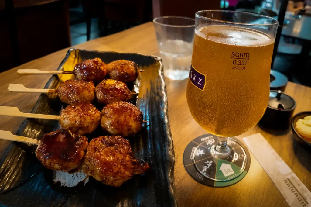 A Japanese izakaya-style meal featuring assorted grilled chicken meatball skewers glazed with savory sauce and arranged on a rectangular black ceramic plate. The skewers are paired with a chilled glass of golden draft beer served in a branded beer glass, with condensation highlighting its refreshing appearance. Set on a wooden table in a cozy restaurant, the meal captures the classic combination of yakitori and beer, creating a warm and inviting casual dining experience.