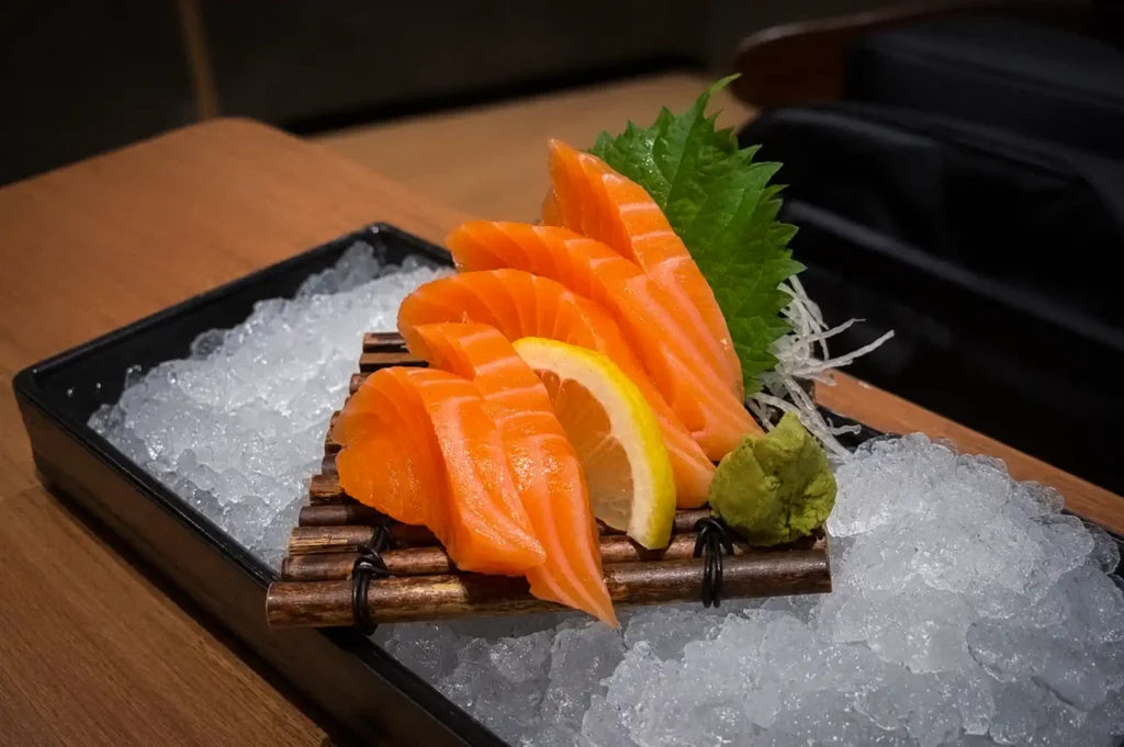 Fresh salmon sashimi served on crushed ice with wasabi, lemon, shredded daikon, and shiso leaf at Issho Izakaya, showcasing authentic Japanese seafood presentation.