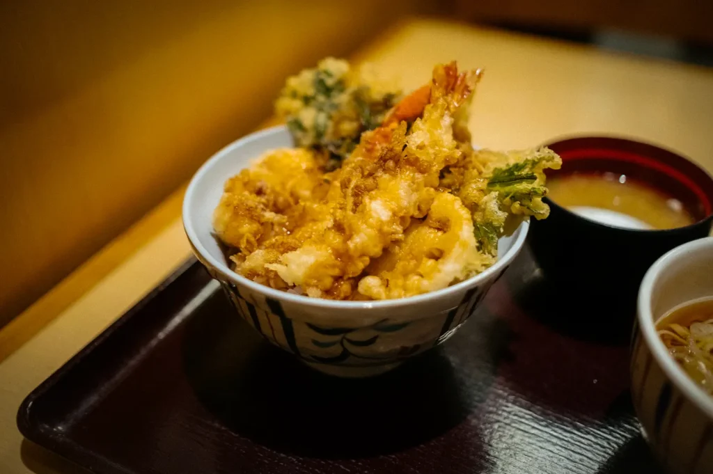 Japanese tendon rice bowl topped with crispy shrimp and vegetable tempura, served with miso soup and noodles on a traditional meal tray.