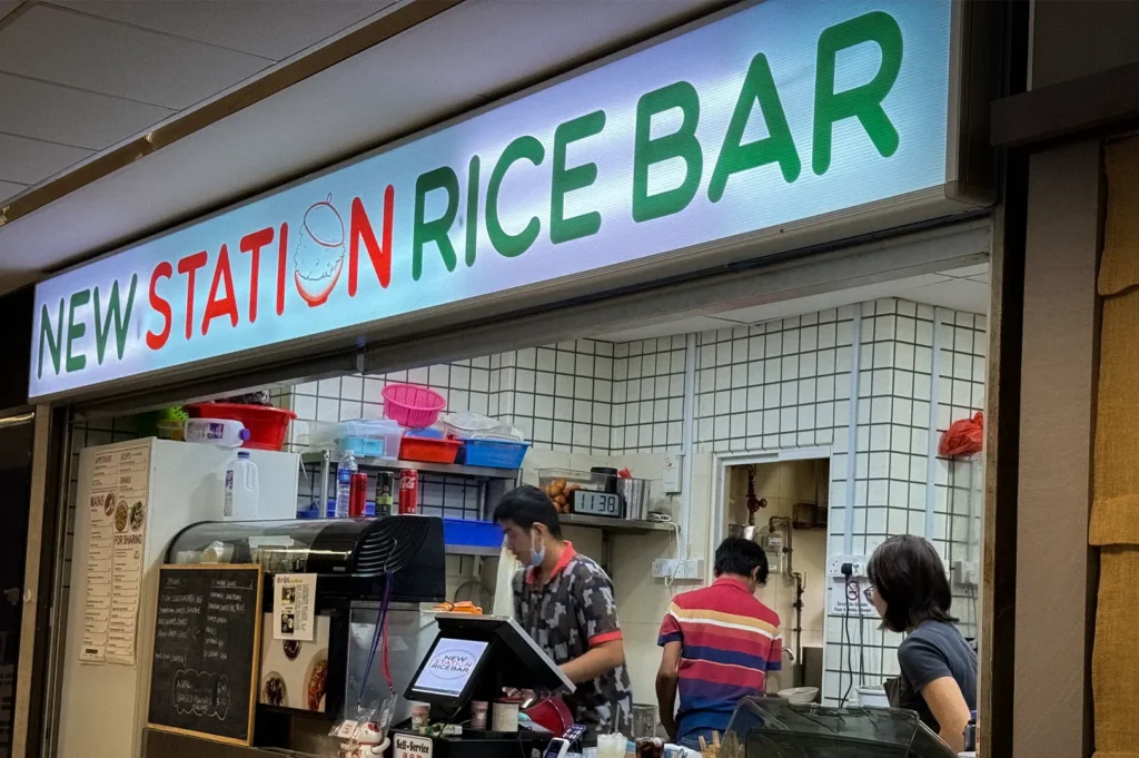 Exterior of New Station Rice Bar in Singapore featuring the restaurant signage, open kitchen, service counter, and staff preparing meals inside.