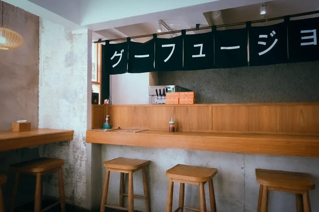 Minimalist Japanese izakaya interior with wooden counter seating, traditional noren curtains, rustic concrete walls, and a warm, inviting dining atmosphere.